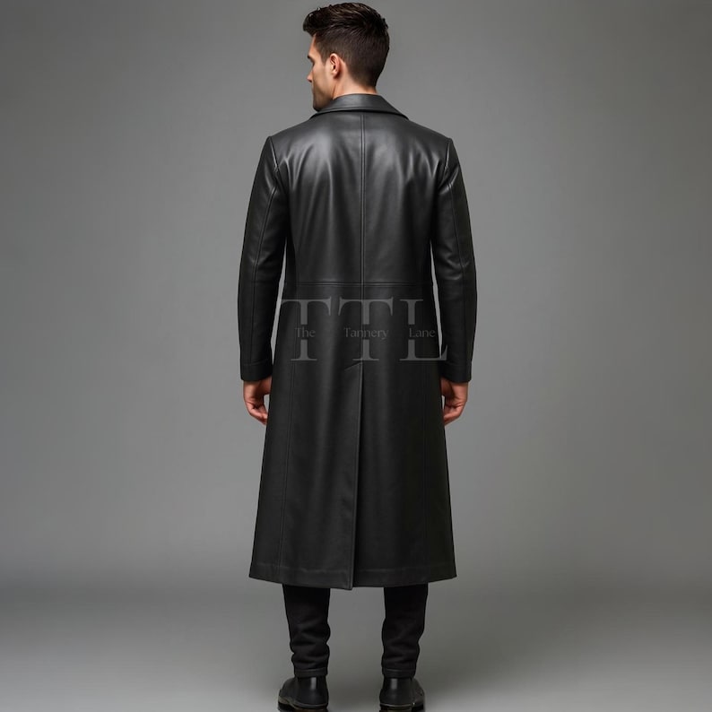 May include: A full-length black leather coat with a notched collar. The coat has a tailored fit and a long, flowing design. The back of the coat features a vent at the hem. The coat is worn with black pants and shoes. The Tannery Lane logo is visible.