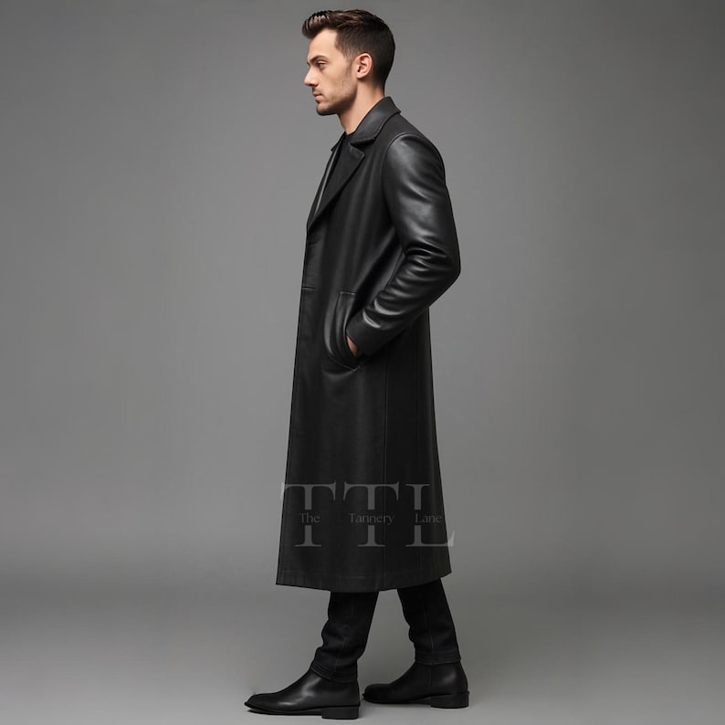 May include: A long, black leather coat with a classic design. The coat features a notched collar, front pockets, and a long length, reaching the ankles. The coat is worn with black pants and shoes. The Tannery Lane logo is visible on the coat.