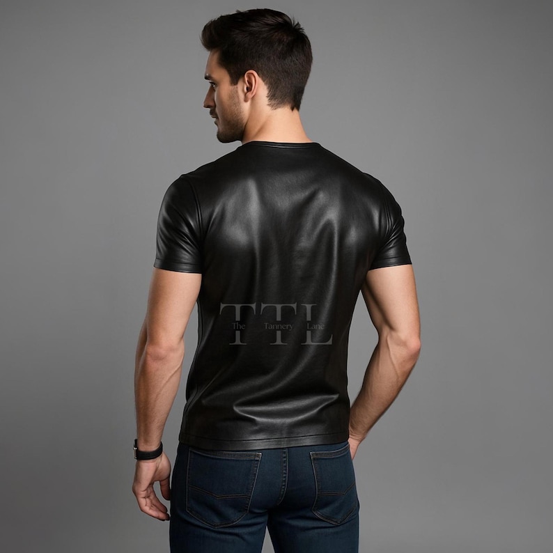 Puede incluir: Camiseta de cuero negra con mangas cortas. La camiseta tiene cuello redondo y un brillo sutil. El texto "The Tannery Lane" est&aacute; impreso en la parte posterior de la camiseta. El modelo lleva vaqueros azul oscuro y un reloj negro.