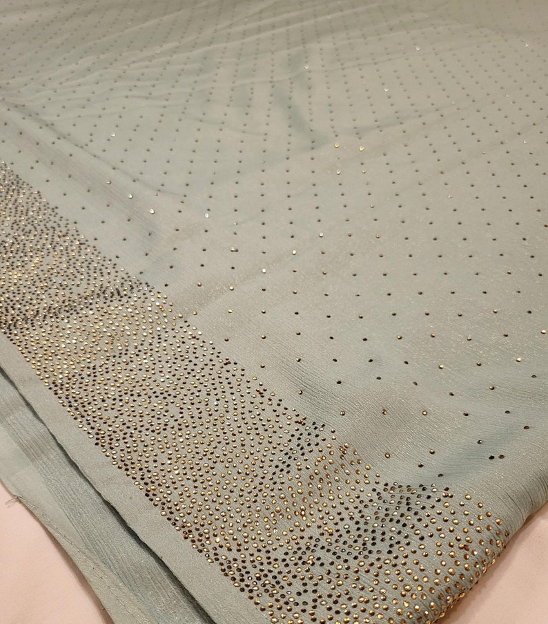 May include: A light blue fabric with a pattern of small gold sequins. The sequins are arranged in a repeating pattern, creating a subtle shimmer effect. The fabric is soft and smooth to the touch.