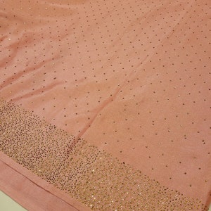 May include: A blush pink fabric with a subtle sheen, densely embellished with small, gold-colored rhinestones along the bottom edge. Scattered rhinestones dot the rest of the fabric. The fabric is folded, showing a layered effect.