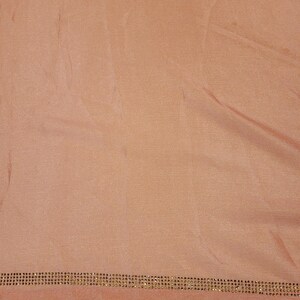 May include: A peach-colored fabric with a subtle sheen. The bottom edge is embellished with a row of small, evenly spaced, gold-colored rhinestones. The fabric is slightly wrinkled, suggesting it is draped or folded.
