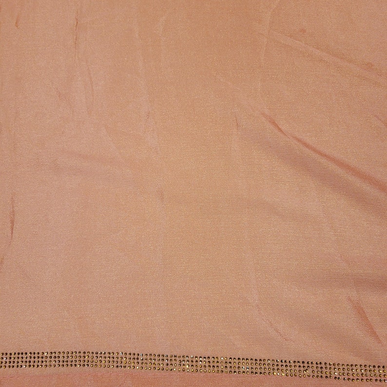 May include: A peach-colored fabric with a subtle sheen. The bottom edge is embellished with a row of small, evenly spaced, gold-colored rhinestones. The fabric is slightly wrinkled, suggesting it is draped or folded.