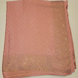 May include: A blush pink scarf with a border and scattered accents of small, sparkling embellishments. The scarf is folded, showcasing the texture and design. The embellishments are a warm gold color.