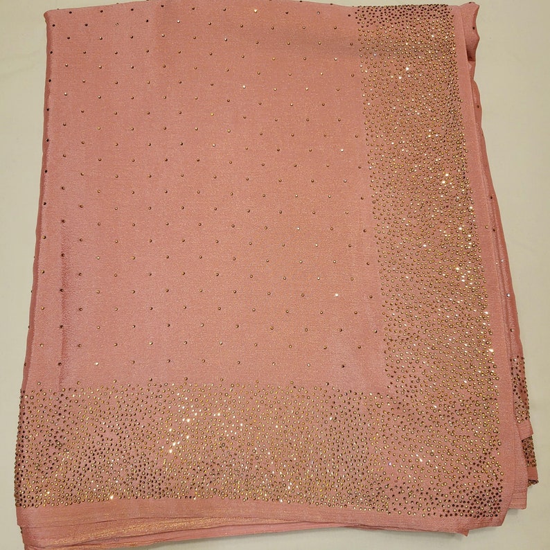 May include: A blush pink scarf with a border and scattered accents of small, sparkling embellishments. The scarf is folded, showcasing the texture and design. The embellishments are a warm gold color.