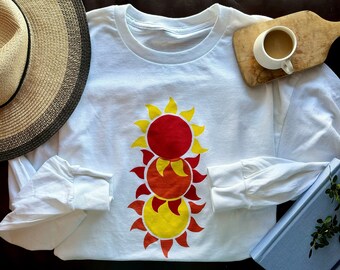 May include: White long-sleeve t-shirt with a graphic of three stacked suns. The suns are red, orange, and yellow with a red outline.