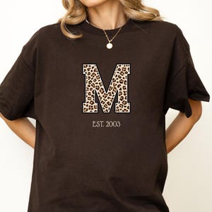Könnte beinhalten: Braunes T-Shirt mit einem großen Buchstaben "M" im Leopardenmuster. Der Text "EST. 2003" ist darunter gedruckt. Das T-Shirt hat eine lockere Passform.