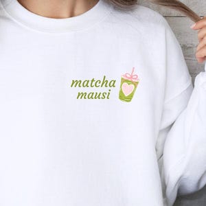 Könnte beinhalten: Weißes Sweatshirt mit dem grünen Schriftzug "matcha mausi". Eine kleine Grafik eines Matcha-Getränks mit rosa Schleife und Herz befindet sich auf der rechten Seite. Das Sweatshirt hat einen lässigen Stil.