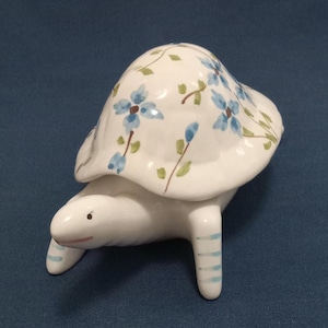 May include: A white ceramic turtle figurine with blue and green floral designs on its shell. The turtle has a closed mouth and blue and white striped legs.