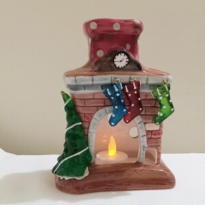 May include: A ceramic fireplace figurine with a red chimney, brick facade, and a lit candle inside. There are three stockings hanging from the mantelpiece, and a small green Christmas tree on the side.