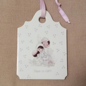 May include: A white ceramic ornament with a pink ribbon. The ornament features a picture of three children holding hands and the text "Peace on Earth".