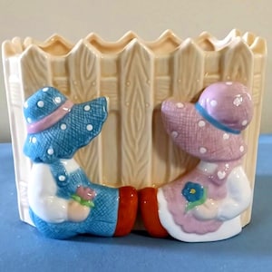 May include: A cream-colored ceramic planter shaped like a picket fence. Two figures are attached to the front, one wearing a blue polka-dot hat and blue overalls, the other wearing a pink polka-dot hat and a pink dress, both holding small flowers.