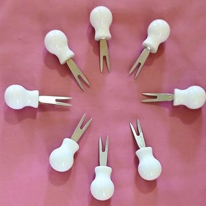 May include: A collection of eight white-handled appetizer forks arranged in a circular pattern on a pink surface. Each fork has a rounded white handle and a two-pronged, silver-colored fork.