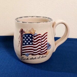 May include: A cream-colored ceramic mug with a distressed rim, featuring a graphic of a waving American flag, small stars, and hearts. The text "Land that I love" is printed in black script below the flag design on the front of the mug.