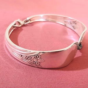 May include: A silver bracelet crafted from repurposed silverware. The bracelet features a floral design and a clasp closure. The bracelet is displayed on a pink surface.