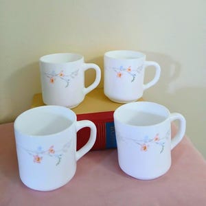 May include: A set of four white milk glass mugs, each featuring a delicate floral pattern with small orange and blue blossoms on thin green stems. The mugs have a smooth, cylindrical shape with a curved handle on the side, arranged in a group.