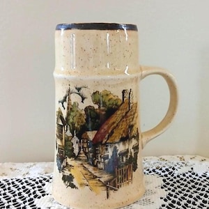 May include: A tall, cylindrical ceramic beer mug with a speckled cream-colored glaze and a dark brown rim. The front features a detailed, colorful illustration of a village scene with thatched-roof cottages, trees, and a church spire along a path.