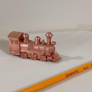 May include: A small, copper-colored toy train with detailed features, including wheels, a smokestack, and a cab. The train is positioned on a white surface next to a yellow pencil with the text "GRAPHITE No.2 HB".