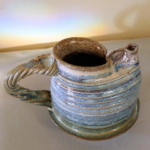 May include: A ceramic pitcher featuring a blue and brown glaze with horizontal ribbed textures. The vessel has a wide, open mouth, a small pouring spout, and a thick, twisted handle that extends from the side of the rounded body.