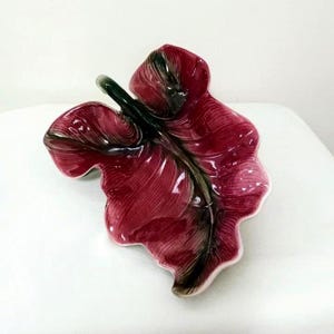 May include: A ceramic leaf-shaped dish in shades of burgundy and dark green. The dish has a glossy finish and detailed veining, with a light pink rim. The decorative piece is designed to resemble a leaf.