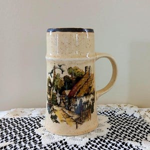 May include: A tall, beige ceramic mug with a handle, featuring a detailed village scene. The mug has a dark brown rim and a speckled texture. The village scene includes houses, trees, and a pathway, painted in muted tones. The mug is likely for beverages.
