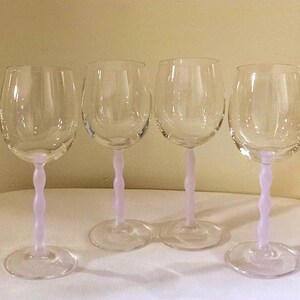 May include: Four clear wine glasses with a light purple, twisted stem. The glasses are on a white surface, and the background is a light beige color. The glasses are empty and ready to be filled.