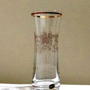 May include: A clear glass vase with a gold rim and a decorative floral pattern. The vase has a slender, slightly flared shape and a small, oval sticker near the base. The gold accents add a touch of elegance to the design.