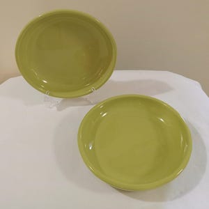 May include: Two lime green ceramic bowls. One bowl is elevated on a clear plastic stand. The bowls have a smooth, glossy finish. The bowls are round with a slightly curved rim. The bowls are empty.