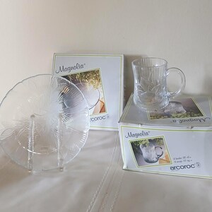 May include: Clear glass plate and mug set with a floral design. The plate is round with a scalloped edge and sits on a clear stand. The mug has a handle and is on top of a box that says "Magnolia" and "arcoroc".