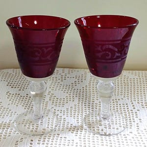 May include: Two wine glasses featuring deep red bowls with a clear, etched scrollwork pattern around the upper section. Each glass has a clear, smooth stem and a circular base, providing a stable foundation for the vibrant red glass vessel.