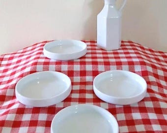May include: A white ceramic oil cruet with a handle and lid, and four small white ceramic bowls are arranged on a red and white checkered tablecloth. The cruet has a geometric shape.