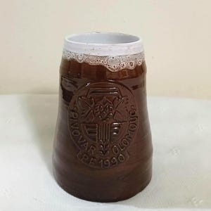 May include: A brown ceramic beer stein with a white rim and a decorative emblem. The stein has a textured surface and a wide base, tapering towards the top. The emblem features text and a design.