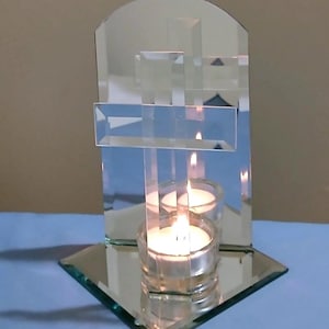 May include: A mirrored candle holder with a cross design. The holder features a clear glass base and a mirrored backdrop with a cross shape. Two lit candles are inside the holder, reflecting in the mirror.