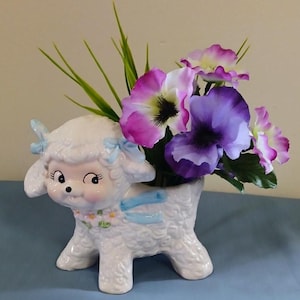 May include: A white ceramic lamb planter featuring a textured wool finish, light blue bows on the ears, and a small floral garland around the neck. The planter is filled with artificial green grass and purple and white pansy flowers.
