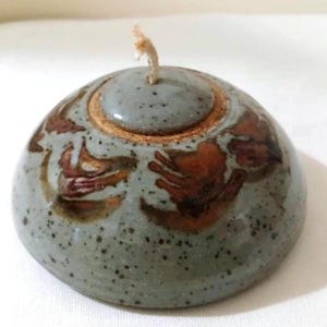 May include: A dome-shaped ceramic oil lamp with a speckled light blue-gray glaze and a central wick. The surface features abstract reddish-brown painted motifs and a circular lid component at the top, showcasing a textured, earthy clay rim.