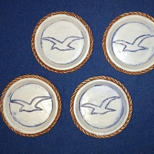 May include: Four round ceramic coasters featuring a white center with a hand-painted blue seagull outline. Each coaster has a textured, rope-like brown border around the edge, creating a nautical appearance for these circular drink accessories.