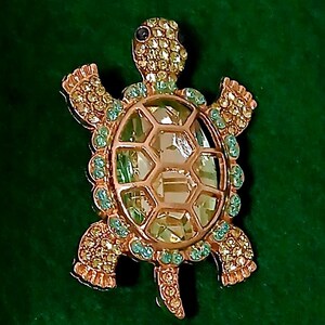 May include: A gold-toned turtle brooch with a large, faceted, light green center. The brooch is encrusted with small, sparkling yellow and light green crystals. The turtle's head features a dark blue crystal eye.
