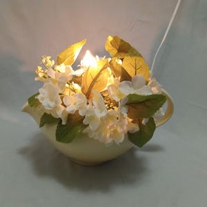 May include: A white ceramic creamer with a light bulb inside, decorated with white flowers and green leaves.