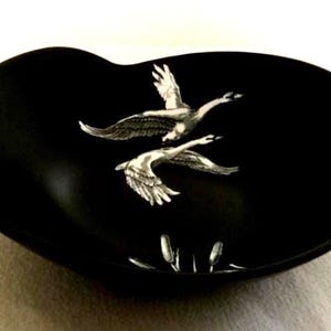May include: Black decorative dish with a unique shape. The dish features two flying geese in a silver color, with reeds at the bottom. The dish is a great addition to any home decor.