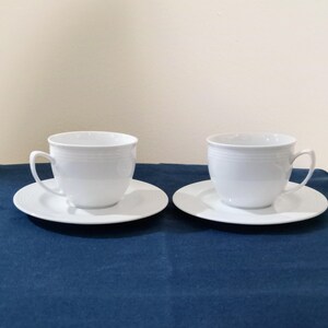 May include: Two white teacups and saucers are displayed on a dark blue surface. The cups have a simple, elegant design with a handle and a slightly flared rim. The saucers are round and have a subtle textured pattern.