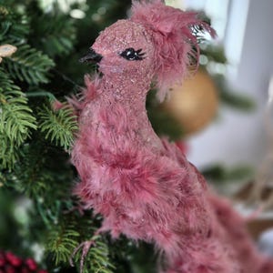 May include: A pink bird ornament with a glittery body and feathery details. The bird has a black beak and is perched on a Christmas tree branch. The ornament is a festive decoration.