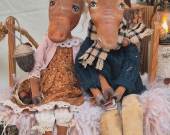 May include: Two handmade moose dolls are seated on a bench. One wears a floral dress, pink cardigan, and a hat. The other wears a blue sweater and a plaid scarf. They are surrounded by decorative elements.