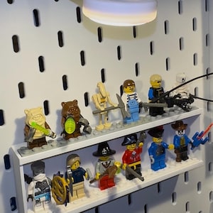 Könnte beinhalten: Eine Sammlung von Lego-Minifiguren, die auf einem weißen Regal ausgestellt und von einer weißen LED-Lampe beleuchtet werden. Die Figuren repräsentieren verschiedene Themen, darunter Piraten, Cowboys und Fantasy-Charaktere. Das Regal ist an einer weißen Lochwand befestigt.