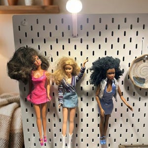 May include: Three fashion dolls are displayed on a white pegboard. The dolls are dressed in various outfits, including a pink dress, a striped top with a denim skirt, and a blue dress with a silver vest. A small light is attached to the pegboard.