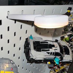 May include: A white LED desk lamp is mounted on a white pegboard. Below the lamp are toy vehicles, including a grey and black futuristic car and a yellow and grey train. The lamp is adjustable.