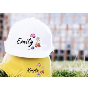 May include: Two construction hard hats, one white and one yellow, stacked on top of each other. The white hard hat has the name "Emily" in black script, along with pink and brown construction-themed graphics. The yellow hat has the name "Krista" and similar graphics.