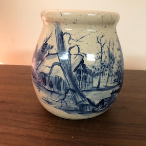 May include: A ceramic jar with a cream-colored base and a blue landscape painting. The jar features a rounded shape with a slightly flared rim. The landscape depicts trees, a house, and a stream, all rendered in shades of blue.
