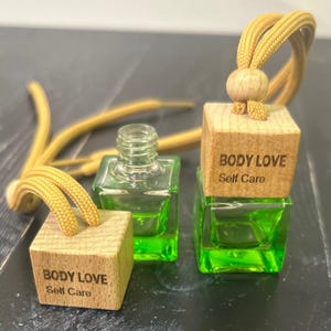 May include: Two small, square glass bottles filled with bright green liquid. Each bottle has a wooden cube top with the words "BODY LOVE Self Care" engraved on it. A yellow cord is attached to the wooden cube, designed for hanging.