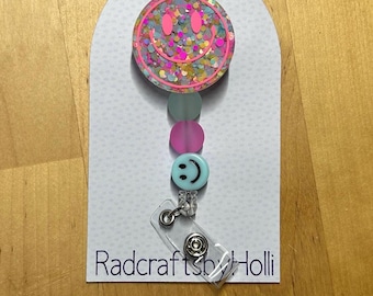 May include: A pink and blue glitter smiley face badge reel with a clear plastic retractable badge holder. The reel has a pink and blue smiley face charm and a clear plastic charm with a black smiley face. The text "Radcrafts by Holli" is printed on the white card.