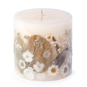 May include: A white candle with dried flowers and leaves embedded in the wax. The candle is in a cylindrical glass jar and has a label that reads "Remarkable Blooms Hand pressed natural floral pillars".
